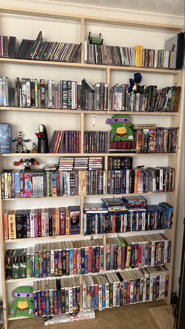 Floor-to-ceiling bookshelf filled with VHS tapes, DVDs, Blu-rays, video games, and CDs